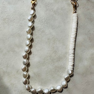 May include: A white phone charm with two gold clasps. One side of the charm is made of white pearls and the other side is made of white shell beads.