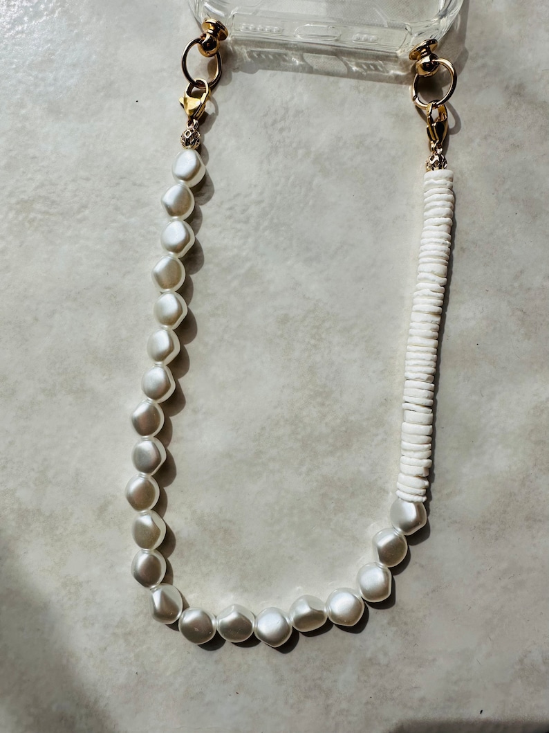 May include: A white phone charm with two gold clasps. One side of the charm is made of white pearls and the other side is made of white shell beads.