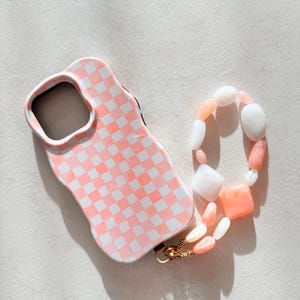 May include: A pink and white chequered phone case with a wavy edge, connected to a beaded strap. The strap features white, pink, and peach beads, attached with a gold-coloured clasp.