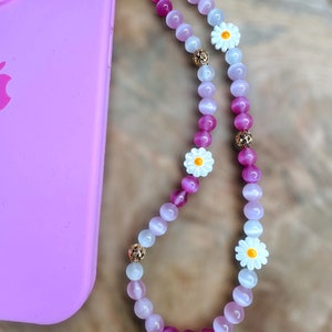 May include: A pink and white beaded phone strap with white daisy charms. The strap is attached to a pink phone case.