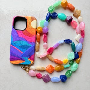 May include: A colorful phone case with a landscape design featuring a sun and mountains, paired with a beaded phone strap. The strap includes a variety of colorful beads in different shapes and sizes, including pink, blue, and orange.