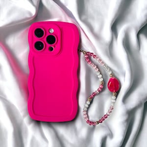 以下が含まれることがあります： 波状の縁が付いた明るいピンク色の携帯電話ケース。 ピンク、白、ゴールドの色合いのビーズの携帯電話チャームが取り付けられています。 チャームには大きなピンク色のペンダントが付いています。