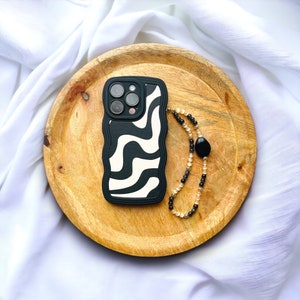 Peut inclure: Une coque de téléphone à motif noir et blanc avec une dragonne à perles. La coque de téléphone a un design ondulé et la dragonne a des perles noires et beiges.
