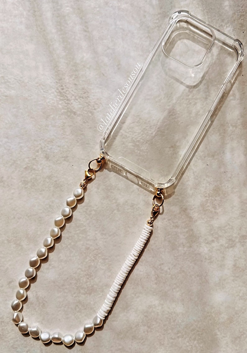 May include: A clear phone case with a gold chain and a white pearl and white bead phone strap. The strap is attached to the phone case with gold clasps.