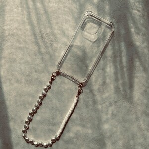 May include: A clear phone case with a white and pearl beaded phone strap. The strap is attached to the case with gold clasps.
