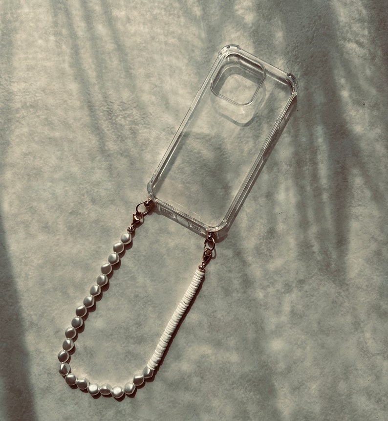 May include: A clear phone case with a white and pearl beaded phone strap. The strap is attached to the case with gold clasps.