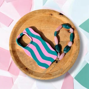 Puede incluir: Una funda de teléfono a rayas rosas y turquesas descansa sobre una bandeja de madera, acompañada de un collar de cuentas con piedras rosas y verdes. La funda tiene un diseño ondulado. El collar tiene un cierre dorado.