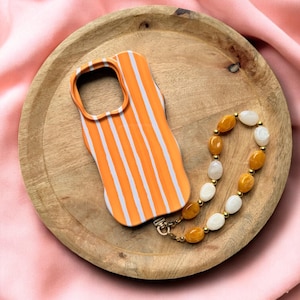 Könnte beinhalten: Orange-weiß gestreiftes Handy-Case mit einer goldenen Kette und einem Perlen-Handyband mit weißen und orangefarbenen Perlen.