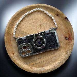 May include: A black and silver phone case designed to resemble a vintage camera, with the text "4800W" and a pearl strap. The case is on a wooden plate against a black background.
