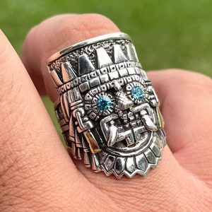 May include: A silver ring with a detailed design of a face with blue gemstones for eyes. The ring is worn on a person's finger.