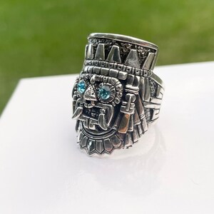 May include: A silver ring with a detailed design of a face with blue gemstones for eyes. The ring is intricately carved and has a textured surface.
