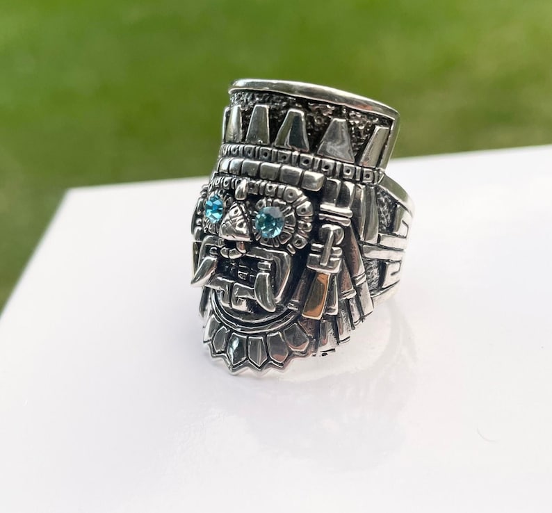 May include: A silver ring with a detailed design of a face with blue gemstones for eyes. The ring is intricately carved and has a textured surface.