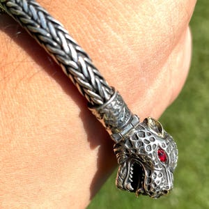 May include: A silver bracelet with a detailed animal head charm. The charm has a red gemstone eye and a textured surface.