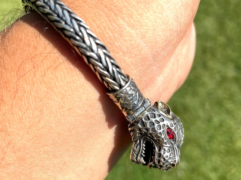 May include: A silver bracelet with a detailed animal head charm. The charm has a red gemstone eye and a textured surface.