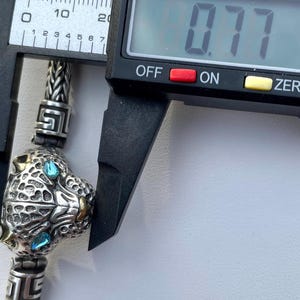 May include: Close-up of a silver charm with a detailed panther head design. The panther has blue eyes and gold accents. The charm is being measured with a caliper, which reads 0.77 inches.