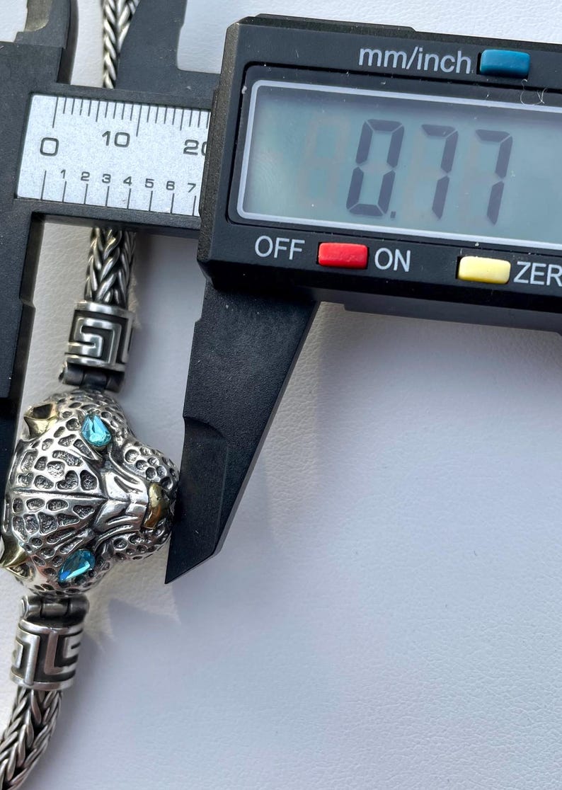 May include: Close-up of a silver charm with a detailed panther head design. The panther has blue eyes and gold accents. The charm is being measured with a caliper, which reads 0.77 inches.