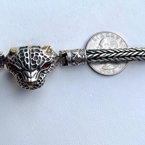 May include: A silver bracelet with a detailed silver and gold panther head charm. The bracelet is made of a braided silver chain.