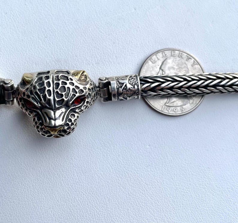 May include: A silver bracelet with a detailed silver and gold panther head charm. The bracelet is made of a braided silver chain.