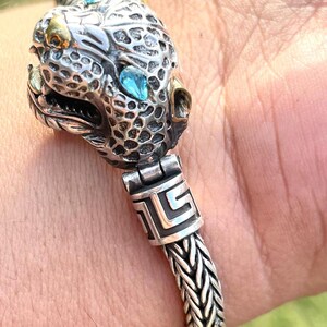 May include: A silver bracelet with a detailed animal head charm featuring blue gemstones. The bracelet has a braided chain design and a decorative clasp.