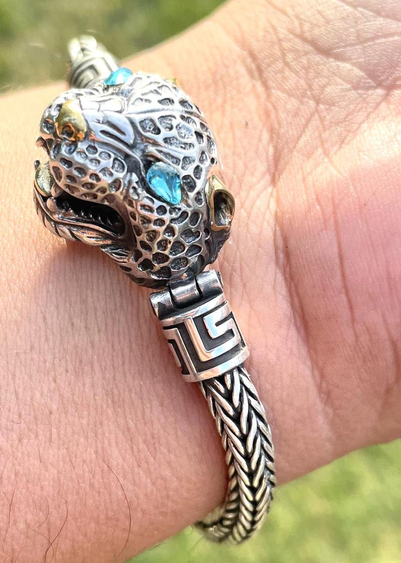 May include: A silver bracelet with a detailed animal head charm featuring blue gemstones. The bracelet has a braided chain design and a decorative clasp.