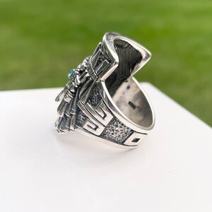May include: Silver ring with a detailed design featuring a turquoise stone. The ring has a textured band and a detailed design on the top.