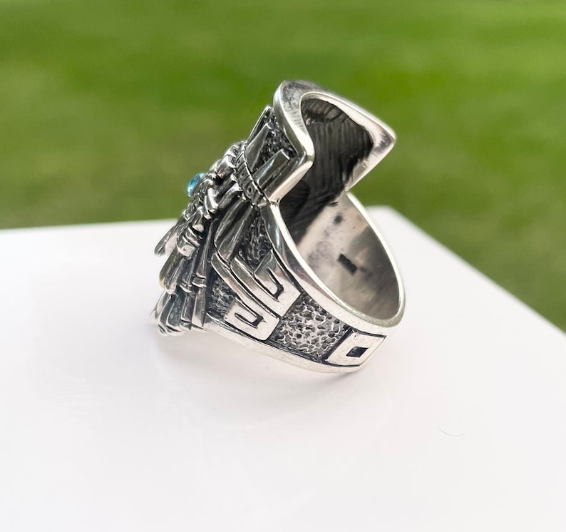 May include: Silver ring with a detailed design featuring a turquoise stone. The ring has a textured band and a detailed design on the top.