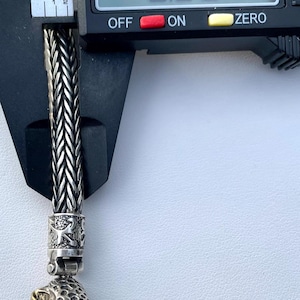 May include: Close-up of a silver chain bracelet with a detailed jaguar head charm. The chain is braided and the charm has red eyes and gold accents. The bracelet is measured with a caliper, showing a measurement of 0.26 inches.