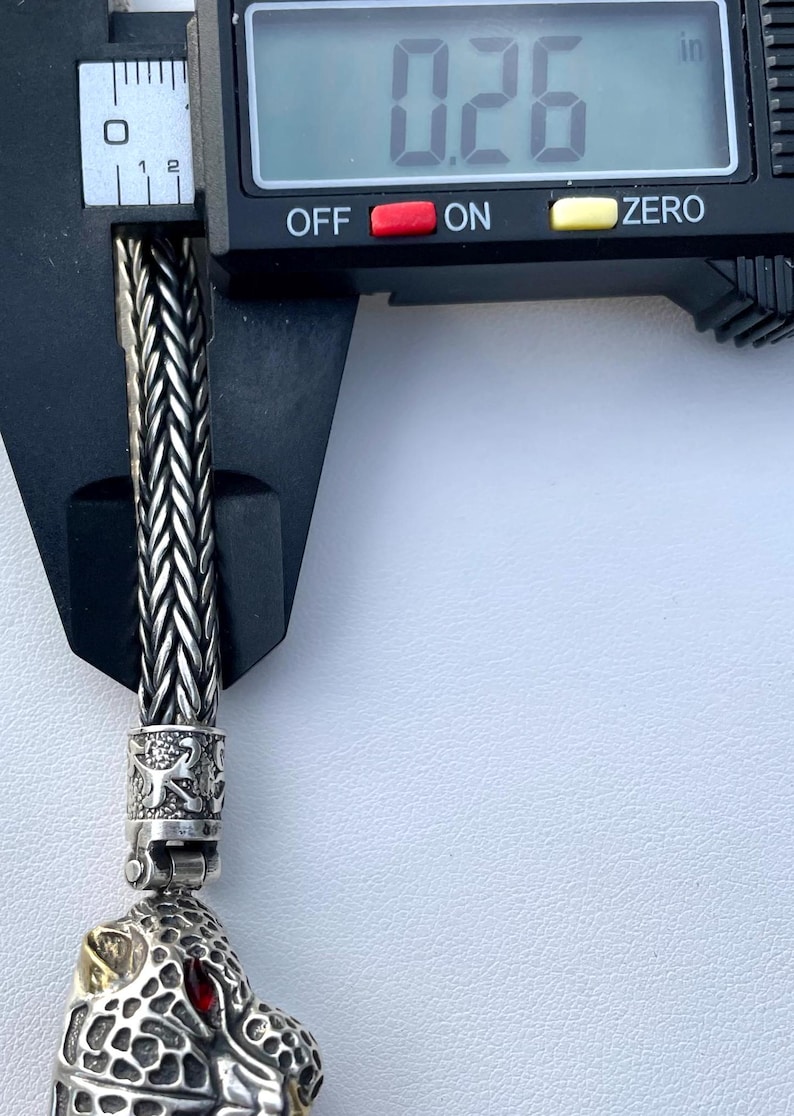 May include: Close-up of a silver chain bracelet with a detailed jaguar head charm. The chain is braided and the charm has red eyes and gold accents. The bracelet is measured with a caliper, showing a measurement of 0.26 inches.
