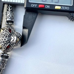 May include: Close-up of a silver chain bracelet with a detailed panther head charm. The charm has red eyes and gold accents. The bracelet is being measured with a caliper, which reads 0.79 inches.