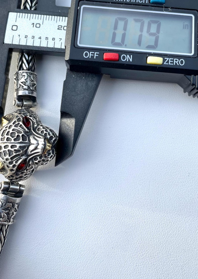 May include: Close-up of a silver chain bracelet with a detailed panther head charm. The charm has red eyes and gold accents. The bracelet is being measured with a caliper, which reads 0.79 inches.