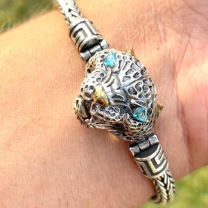 May include: A silver bracelet with a detailed panther head charm featuring two blue gemstones. The bracelet has a braided chain design and a Greek key pattern on the clasp.