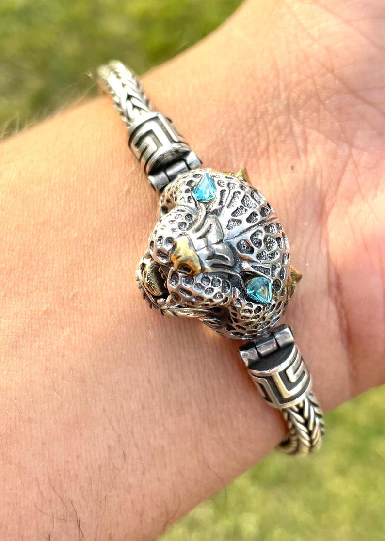 May include: A silver bracelet with a detailed panther head charm featuring two blue gemstones. The bracelet has a braided chain design and a Greek key pattern on the clasp.