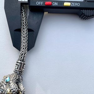 May include: A silver chain bracelet with a detailed silver charm featuring a panther head with blue gemstones for eyes. The chain is 0.22 inches wide.