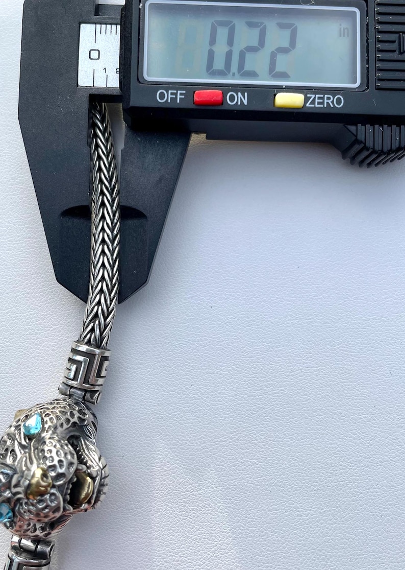 May include: A silver chain bracelet with a detailed silver charm featuring a panther head with blue gemstones for eyes. The chain is 0.22 inches wide.