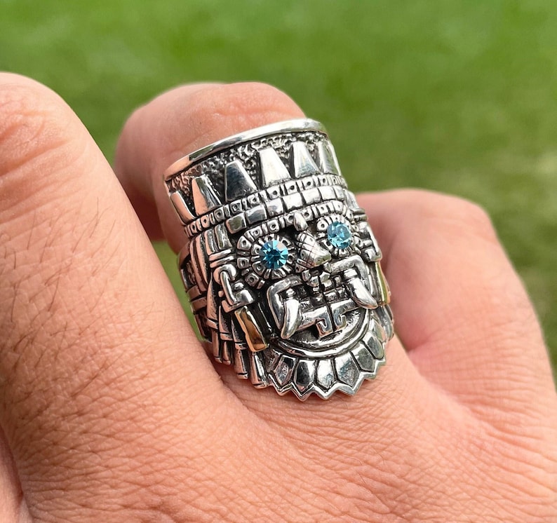 May include: A silver ring with a detailed design of a face with blue gemstones for eyes. The ring is worn on a person's finger.