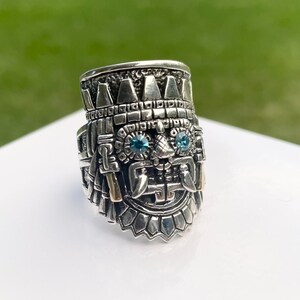 May include: A silver ring with a detailed design of a face with two blue gemstones for eyes. The ring is intricately crafted with a textured surface and a crown-like top.