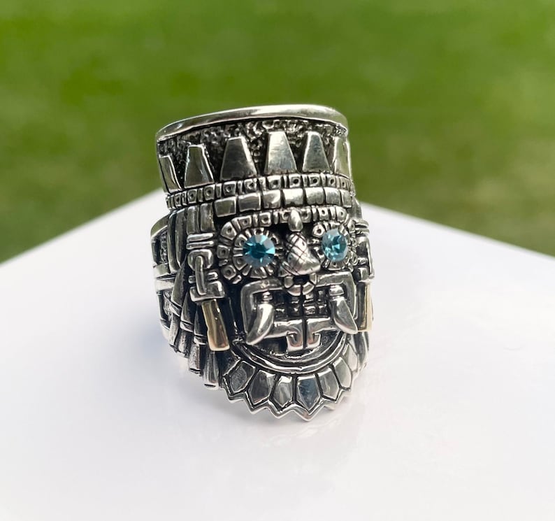 May include: A silver ring with a detailed design of a face with two blue gemstones for eyes. The ring is intricately crafted with a textured surface and a crown-like top.