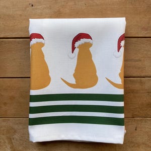 Pode incluir: Toalha de cozinha branca dobrada com um padr&atilde;o repetido de silhuetas de c&atilde;es amarelos usando chap&eacute;us de Papai Noel vermelhos. A parte inferior da toalha tem tr&ecirc;s listras horizontais verdes.
