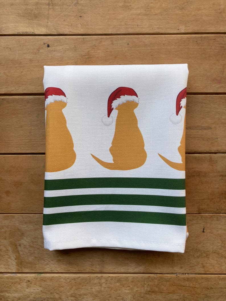 Pode incluir: Toalha de cozinha branca dobrada com um padr&atilde;o repetido de silhuetas de c&atilde;es amarelos usando chap&eacute;us de Papai Noel vermelhos. A parte inferior da toalha tem tr&ecirc;s listras horizontais verdes.