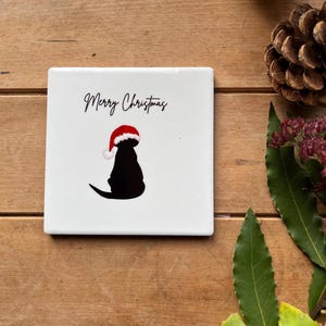 Op de afbeelding: Vierkante witte keramische onderzetter met een zwart silhouet van een hond met een kerstmuts. De woorden "Merry Christmas" staan in zwart script boven de hond. De onderzetter ligt op een houten oppervlak.