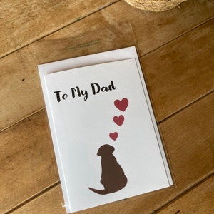 Pode incluir: Um cart&atilde;o branco com o texto "To My Dad" em escrita cursiva preta. Uma silhueta marrom de um cachorro olhando para cima para cinco cora&ccedil;&otilde;es vermelhos.