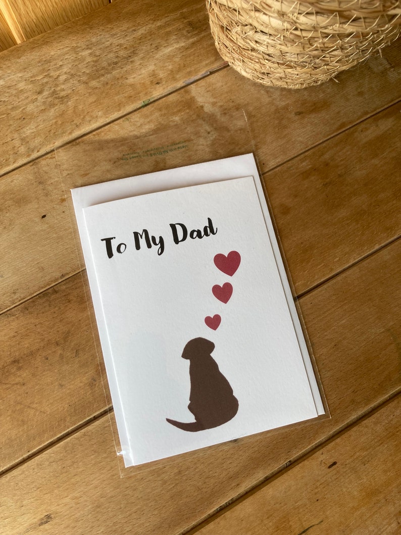 Pode incluir: Um cart&atilde;o branco com o texto "To My Dad" em escrita cursiva preta. Uma silhueta marrom de um cachorro olhando para cima para cinco cora&ccedil;&otilde;es vermelhos.