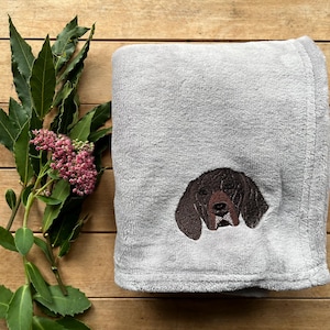 May include: A folded grey blanket featuring an embroidered brown dog's head. The blanket is placed on a wooden surface, accompanied by green leaves and pink flowers.