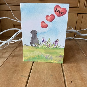 May include: A watercolor greeting card featuring a black dog sitting in a field of flowers. Three red hearts float above the dog, with the word "love" written on the top heart.