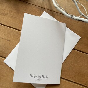 May include: A white card with the text "Madge And Maple" printed in black. The card is on a wooden surface.