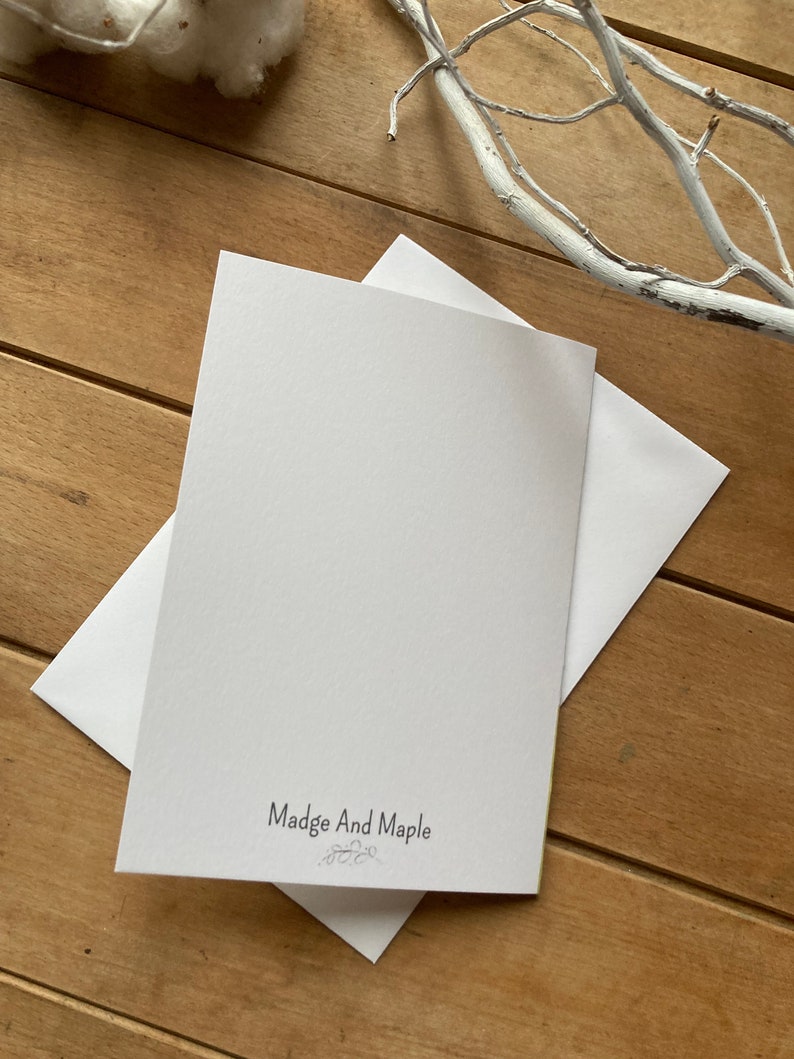 May include: A white card with the text "Madge And Maple" printed in black. The card is on a wooden surface.