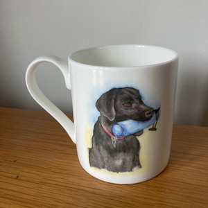 May include: White ceramic mug featuring an illustration of a black Labrador Retriever dog. The dog is holding a blue and white object in its mouth.