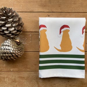 Pode incluir: Toalha de cozinha branca com um padr&atilde;o repetido de silhuetas de c&atilde;es amarelos usando chap&eacute;us de Papai Noel vermelhos. A parte inferior da toalha tem listras verdes e brancas. Dois pinhas est&atilde;o &agrave; esquerda.