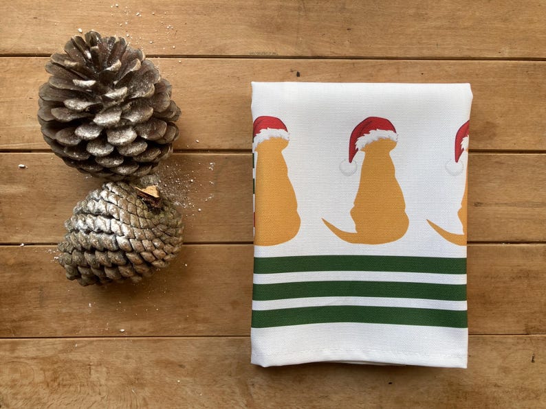 Pode incluir: Toalha de cozinha branca com um padr&atilde;o repetido de silhuetas de c&atilde;es amarelos usando chap&eacute;us de Papai Noel vermelhos. A parte inferior da toalha tem listras verdes e brancas. Dois pinhas est&atilde;o &agrave; esquerda.