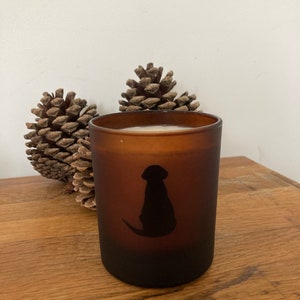 Op de afbeelding: Een bruine, gematteerde glazen kaars met een zwart silhouet van een hond aan de voorkant. De kaars staat op een houten oppervlak met twee dennenappels.